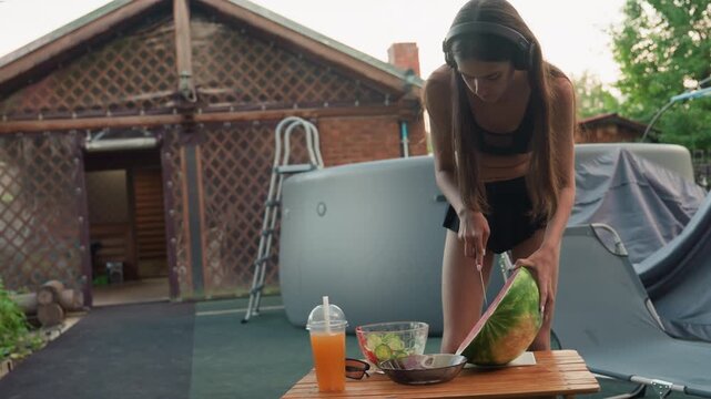 Woman wearing headphones cuts watermelon on outdoor table with juice and salad near camping gear and wooden lattice house in background cutting shorts
