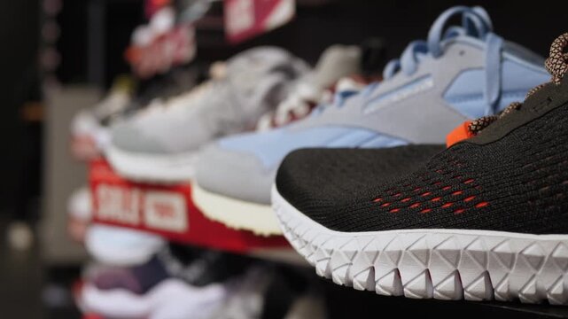 Sneaker display on shop shelf stays in focus while black, blue, and white sports shoes sit in rows behind. Close foreground trainer with textured sole creates sleek retail product showcase mood.