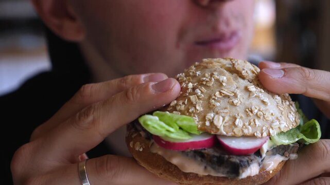 Close up of man eating seeded burger, hands bracing sandwich as he takes repeated bites. Lettuce, radish and creamy sauce show in bun while diner chews with content concentration, fingers gripping