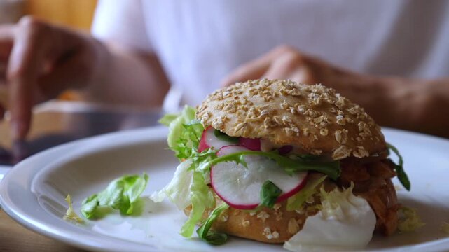 Sesame sandwich rests on white plate as diner hands hover nearby. Crisp vegetables, radish, lettuce and creamy sauce make this plated lunch look fresh, colorful and inviting in soft light.