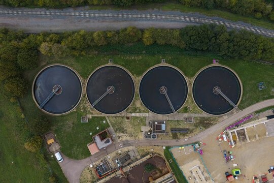 Aerial view of a wastewater treatment plant with four large circular sedimentation tanks surrounded by green fields and a railway line in the United Kingdom.