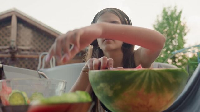 Young woman wearing headphones carefully removes watermelon rind and places fruit into glass bowl with another container of sliced vegetables visible on table. scene takes place outdoors