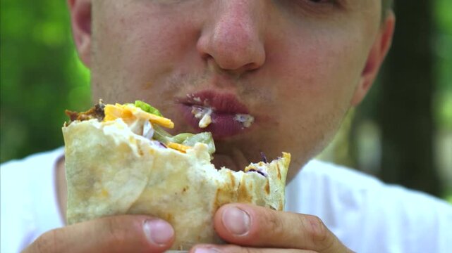 Close up man enjoys burrito outdoors, gripping it tightly while taking large bite. He chews messily and wipes none away, with bright green background adding casual picnic mood. Hands hold wrap firmly.