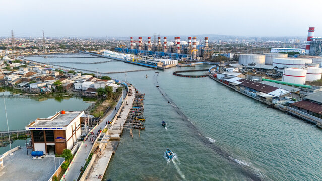Aerial view of a power plant with tall chimneys and storage tanks situated along a water channel with small boats navigating the waterway in Semarang, Central Java, Indonesia.