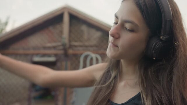Young woman with long hair listens to music through large over ear headphones while sitting outdoors in backyard setting. Woman occasionally moves her arm and background features greenery