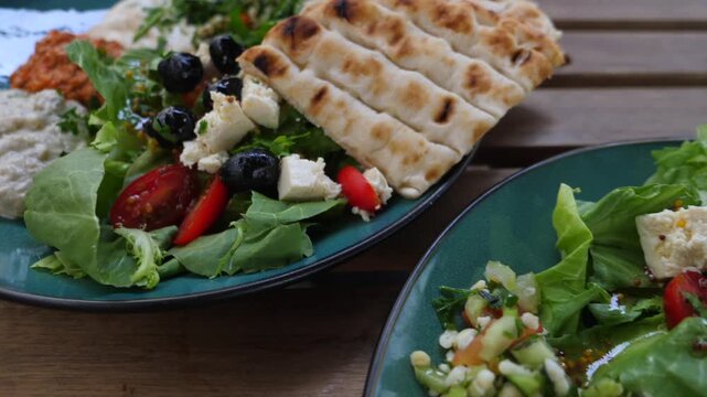 Colorful mezze plate features hummus, eggplant dip, leafy salad, olives and flatbread on rustic table. Mediterranean vegetarian meal appears fresh, hearty and ready to share, with sliced tomato