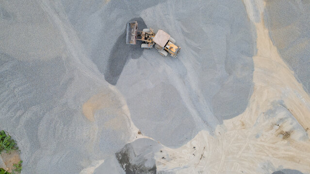 Wheel Loader Surrounded by a Large Aggregate Stockpiling Area in Indonesia. Heavy Construction Machinery Stands Among Rock, Sand, and Processed Materials in Industrial Quarry Operations from Above.