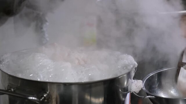 Chef using metal ladle to stir boiling white broth in large industrial kitchen pot cooking soup liquid steam hot cooking soup liquid steam hot