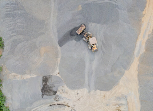 Wheel Loader Surrounded by a Large Aggregate Stockpiling Area in Indonesia. Heavy Construction Machinery Stands Among Rock, Sand, and Processed Materials in Industrial Quarry Operations from Above.