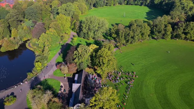 Aerial video looking over the historic pavilion, boating lake, open playing greens, and morning runners in Alexandra Park near Princes Road in Manchester. 