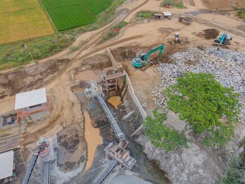 Mining Work in a Rock and Sand Quarry with an Excavator and Wheel Loader Working on a Pile of Large Rocks Near Rock Crusher Machinery. Heavy Equipment Operates Across the Active Mining Site from Above