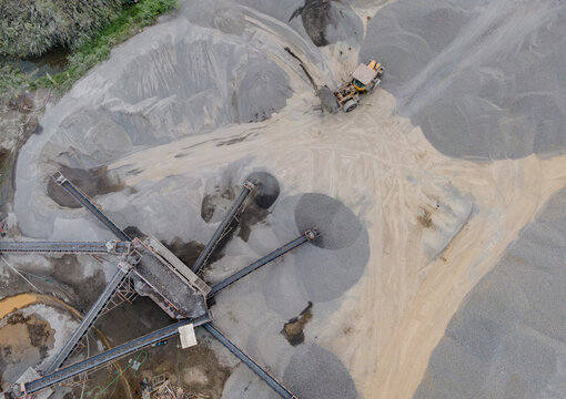 Wheel Loader and Conveyor Belt Transporting Split Rocks in the Middle of an Aggregate Stockpiling Area. Heavy Machinery Operates Among Rock, Sand, and Processed Materials Viewed from Above.