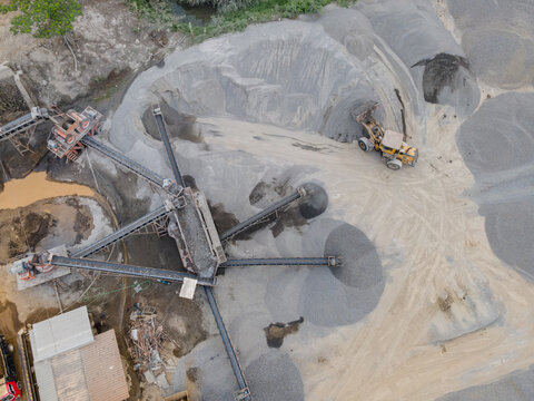 Wheel Loader and Conveyor Belt Transporting Split Rocks in the Middle of an Aggregate Stockpiling Area. Heavy Machinery Operates Among Rock, Sand, and Processed Materials Viewed from Above.