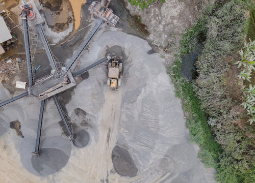 Wheel Loader and Conveyor Belt Transporting Split Rocks in the Middle of an Aggregate Stockpiling Area. Heavy Machinery Operates Among Rock, Sand, and Processed Materials Viewed from Above.