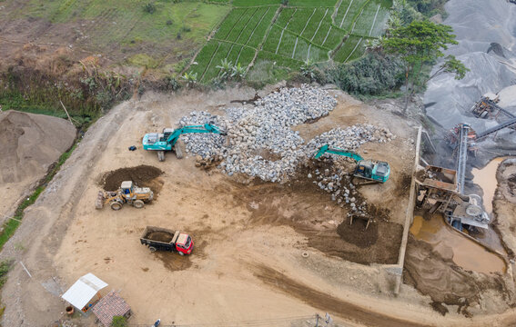 Mining Work in a Rock and Sand Quarry with an Excavator and Wheel Loader Working on a Pile of Large Rocks Near Rock Crusher Machinery. Heavy Equipment Operates Across the Active Mining Site from Above