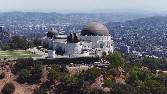 Aerial view shows Griffith Observatory situated on Mount Hollywood in Los Angeles, California, with distant city sprawl under clear daylight as visitors walk along the pathways.
