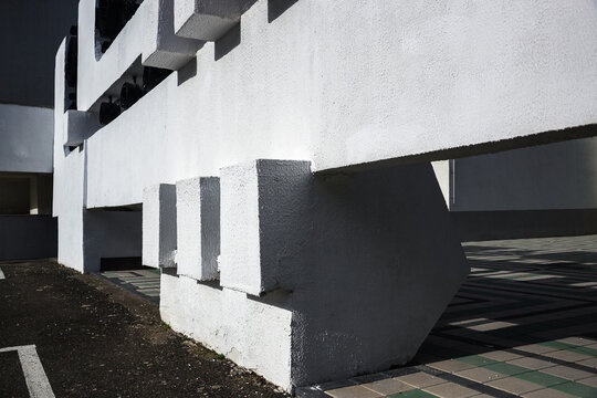 Modern white concrete building exterior with massive geometric structural elements casting harsh abstract shadows on tiled urban plaza ground