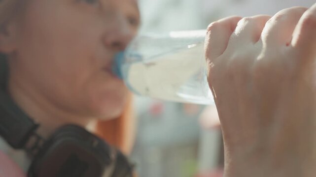 Woman wearing pink shirt and headphones drinks water from clear plastic bottle in bright daylight. focus remains on her hand gripping transparent container capturing subtle movements