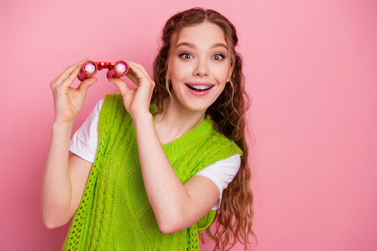 Young girl with pink background smiling holding red binoculars wearing green vest fashion lifestyle