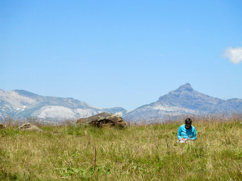 Woman sitting in grassy mountain meadow reading map and writing. A hiker sitting in the high grass of an alpine pasture with rocky mountain peaks in the background, under a clear blue sky.