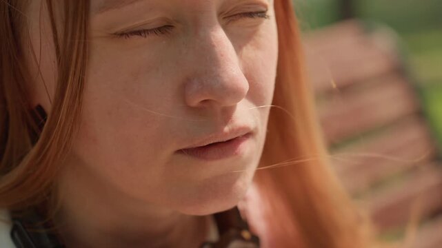 Young woman with long hair holds small object near her mouth possibly applying makeup or using electronic device. scene is set outdoors on sunny day featuring soft natural light and blurred