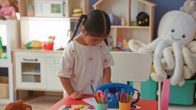 Girl holding blue toy telephone to ear while playing at colorful playroom play table; playful curiosity.