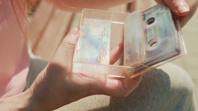 Young woman examines transparent cassette tape case revealing its iridescent plastic and internal components. Woman holds object steadily in her hands against outdoor wooden bench background