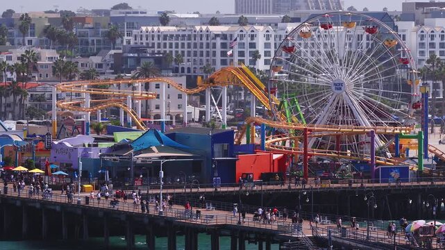 Aerial tracking view reveals Pacific Park rides, a roller coaster, Ferris wheel, shops, and visitors strolling along the historic Santa Monica Pier in California.