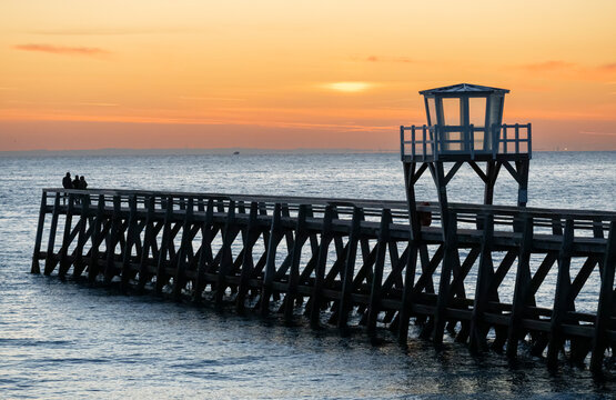 Luc-Sur-Mer, France - 08 08 2026:  View of a wooden pier in front of the sea, a colorful sunrise and port of Le Havre far away