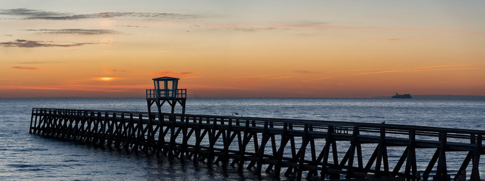 Luc-Sur-Mer, France - 08 08 2026:  View of a wooden pier in front of the sea, a colorful sunrise, a cruise ship and port of Le Havre far behind