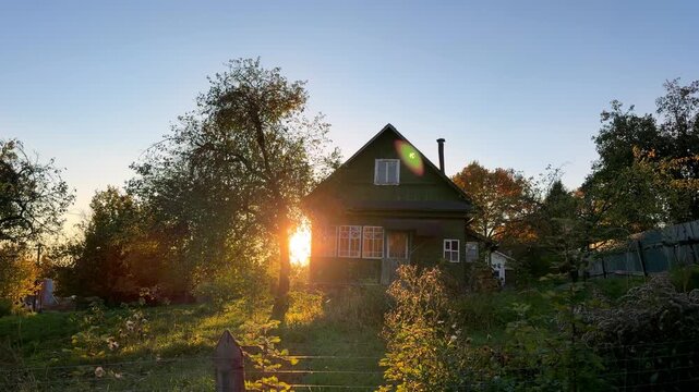 Sunlight moves behind a rustic wooden house as the camera advances slightly, revealing an autumn garden, trees, fence, and clear sky