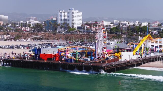 Aerial shot captures the iconic Santa Monica Pier featuring Pacific Park rides, a Ferris wheel, roller coaster, adjacent sandy beach, and the coastal skyline in Los Angeles, California.