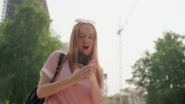 Young woman wearing pink shirt and carrying backpack fidgets with her hands in outdoor urban setting. background features green trees distant buildings including cranes soft daylight