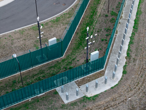 Aerial view of a data center perimeter featuring green security fencing, multiple surveillance cameras mounted on poles, and concrete bollards along a paved walkway in the United Kingdom.