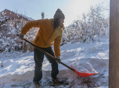 Happy caucasian woman smiling and shoveling snow near a country house terrace on a sunny winter day.