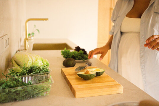 Pregnant woman in a light blue shirt prepares fresh vegetables, including avocado and lettuce, on a wooden cutting board in a modern kitchen setting
