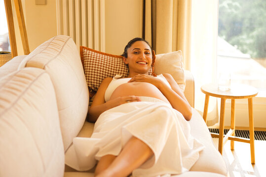 Pregnant woman relaxing on a sofa in a bright living room, wearing a white dress, smiling and gently holding her belly, with natural light streaming through the window