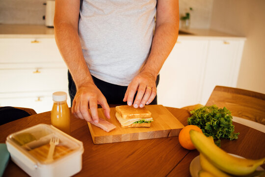 Man in gray t-shirt prepares a sandwich with lettuce and ham on a wooden cutting board in a bright kitchen with fruits and juice on the table
