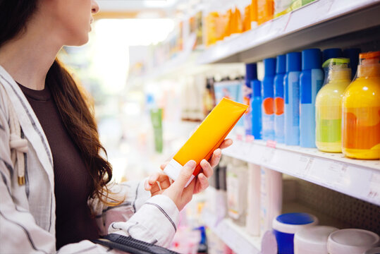 Female shopper examining orange tube of cream in personal care aisle, colorful bottles and containers arranged on shelves in a retail store