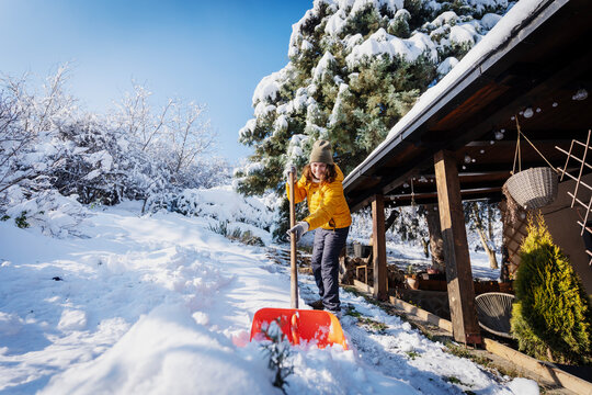 Happy caucasian woman smiling and shoveling snow near a country house terrace on a sunny winter day.