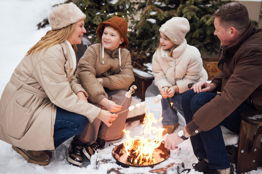 Family enjoying a winter campfire in the snow, roasting marshmallows while wearing warm clothing, surrounded by evergreen trees and a cozy outdoor setting