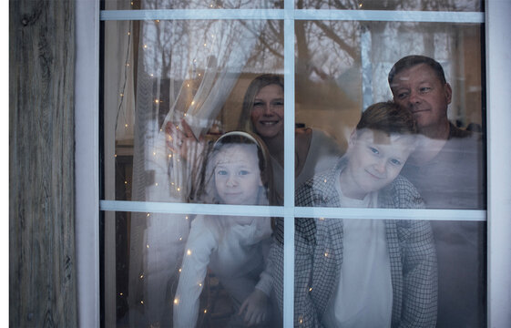 Family of four, including two children, looking through a window with fairy lights, showcasing a cozy indoor atmosphere during winter season