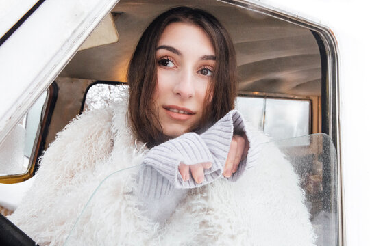 Young woman with long dark hair wearing a cozy white fur coat leans against the window of an old vehicle, showcasing a serene expression in a winter setting
