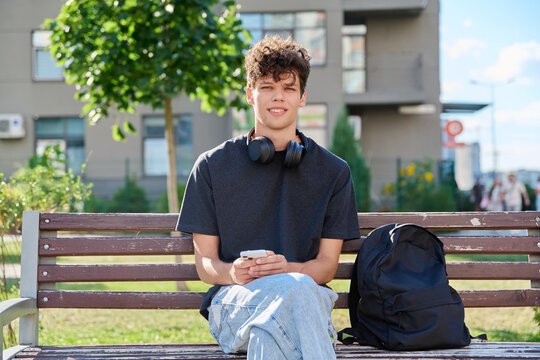Young man with headphones holding smartphone sitting park bench