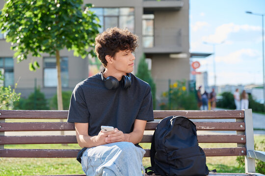Young male student holding smartphone sitting on bench outdoors looking away