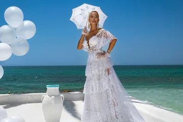 Elegant bride in intricate white lace gown with parasol posing on sunny white terrace with balloons and ancient Greek architecture, overlooking the turquoise Mediterranean ocean © marcink3333