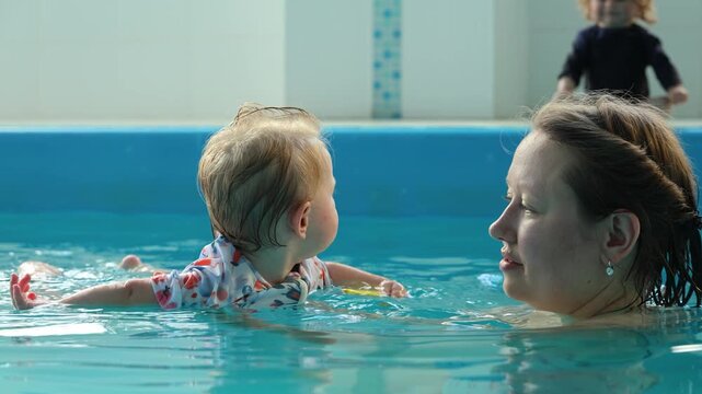 Baby swimming in pool holding duck toy near mother