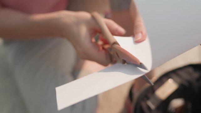 Young woman carefully cuts blank sheet of white paper with pink handled scissors focusing on precise movement and interaction between her hands. scene is set in soft natural light