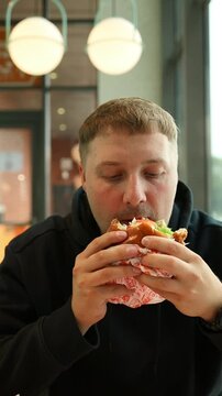 Close-up vertical video portrait of a young caucasian man biting and chewing a large delicious hamburger or sandwich indoors, enjoying fast food. 