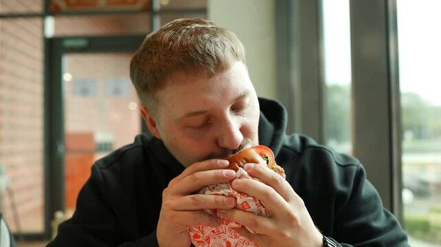 Detailed horizontal shot of a young caucasian man biting and chewing a large delicious hamburger or sandwich indoors, enjoying fast food or a cheat meal.